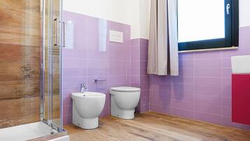 A bathroom with a glass shower enclosure, two toilets, and a window with a curtain.
