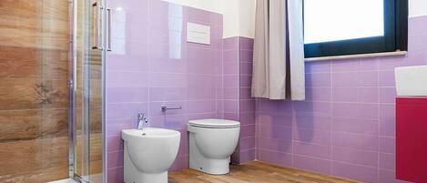 A bathroom with a glass shower enclosure, two toilets, and a window with a curtain.