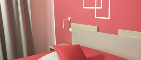 Un lit avec une couvre-lit rouge et un oreiller blanc, une table de chevet avec une lampe, et un mur orné de motifs géométriques.