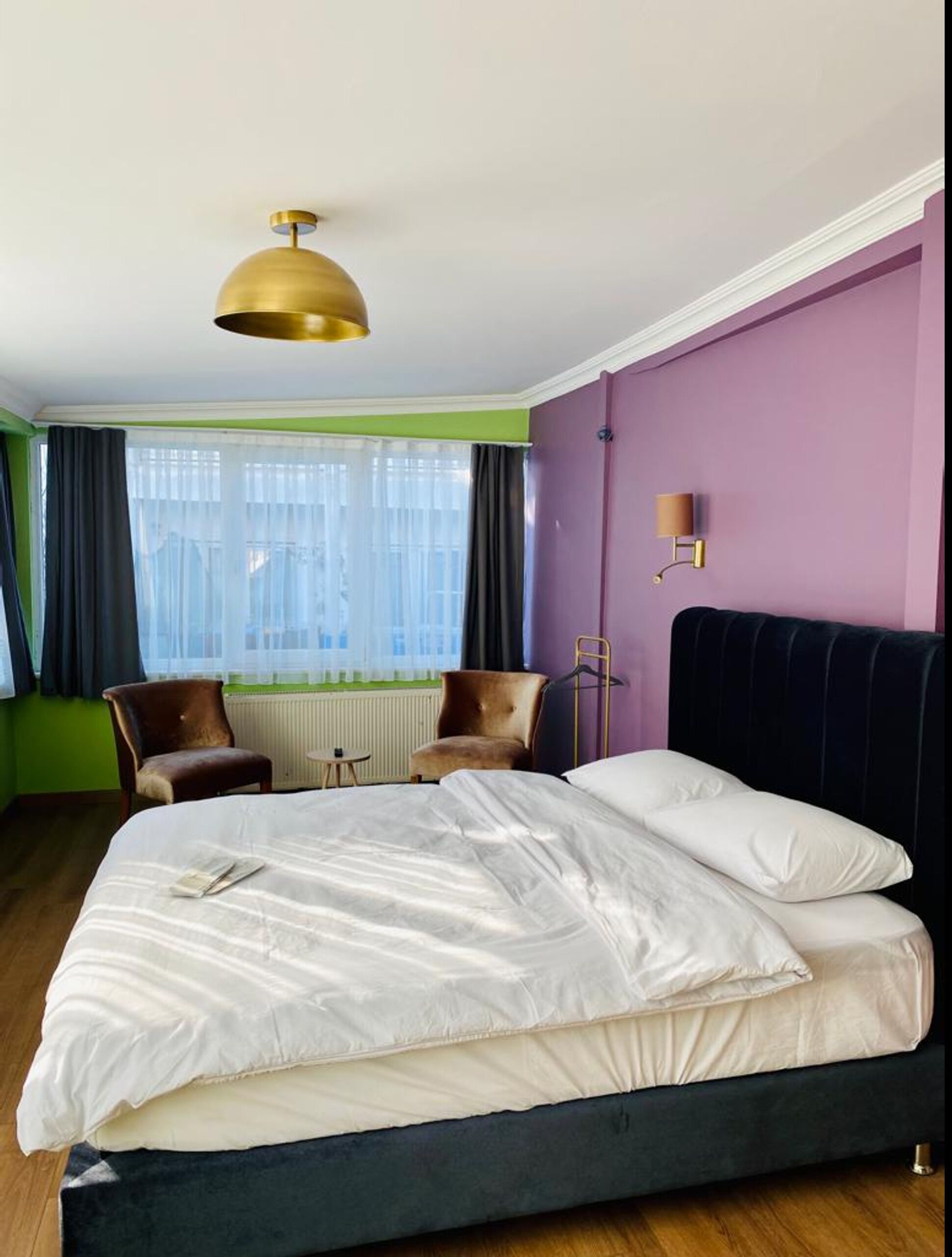 A hotel room with a bed, a chair, a radiator, and a window with curtains.