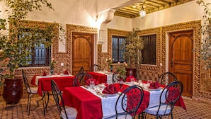 Un comedor con mesas dispuestas para cuatro, cada una con mantel rojo y servilletas blancas. La habitación presenta puertas de madera ornamentadas y piso de baldosas.