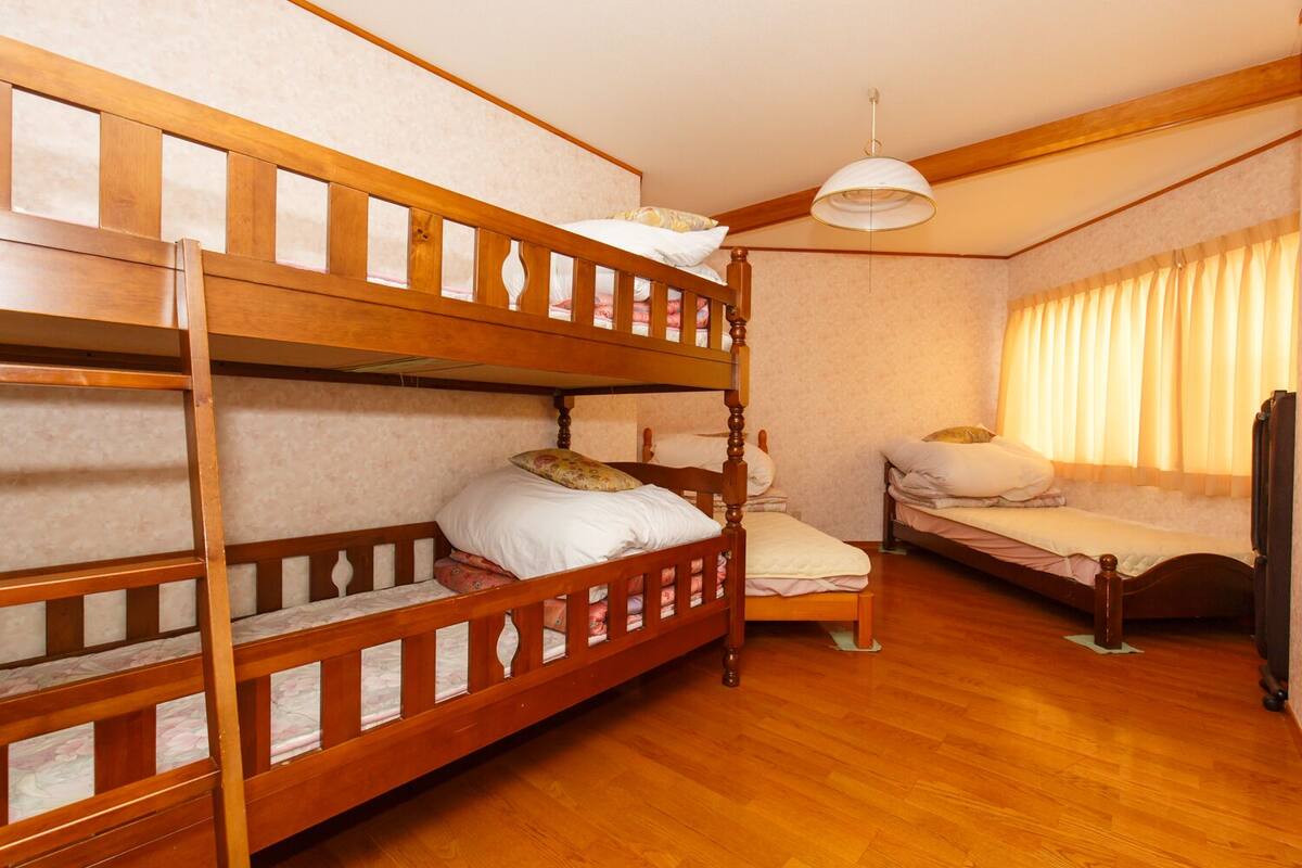 Ein Zimmer mit zwei Etagenbetten und einem Einzelbett, Holzboden, Deckenventilator und einem fenster mit Vorhängen.