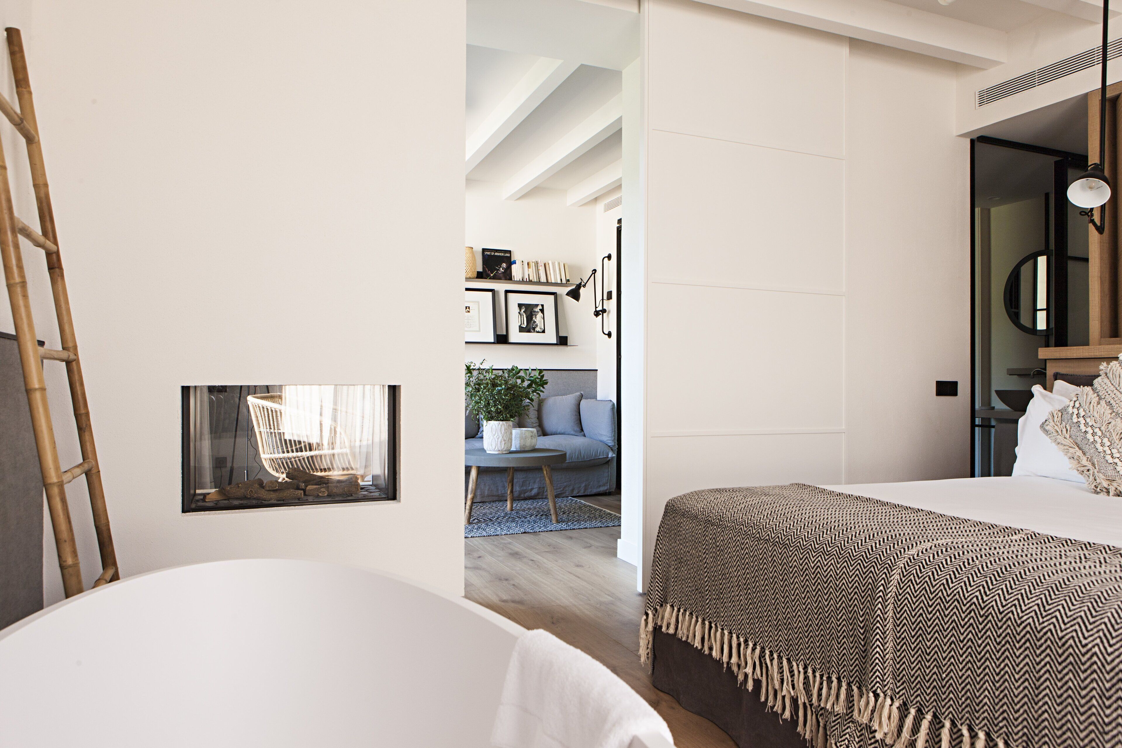 A modern bedroom with a large bed, a bedside table, and a mirror. There is a window with a view of a living room and a staircase.