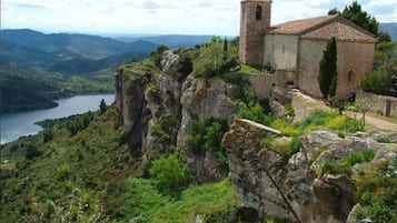 A stone building with a bell tower on a cliff overlooking a valley and a body of water.