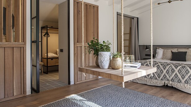 Ein Schlafzimmer mit Hängesessel, einem Bett mit gemusterten Textilien und einem Nachttisch mit Büchern und Pflanzen.