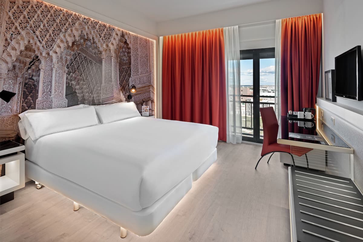 A modern hotel room with a large bed, a desk with a flat-screen TV, and a view of the city through large windows.