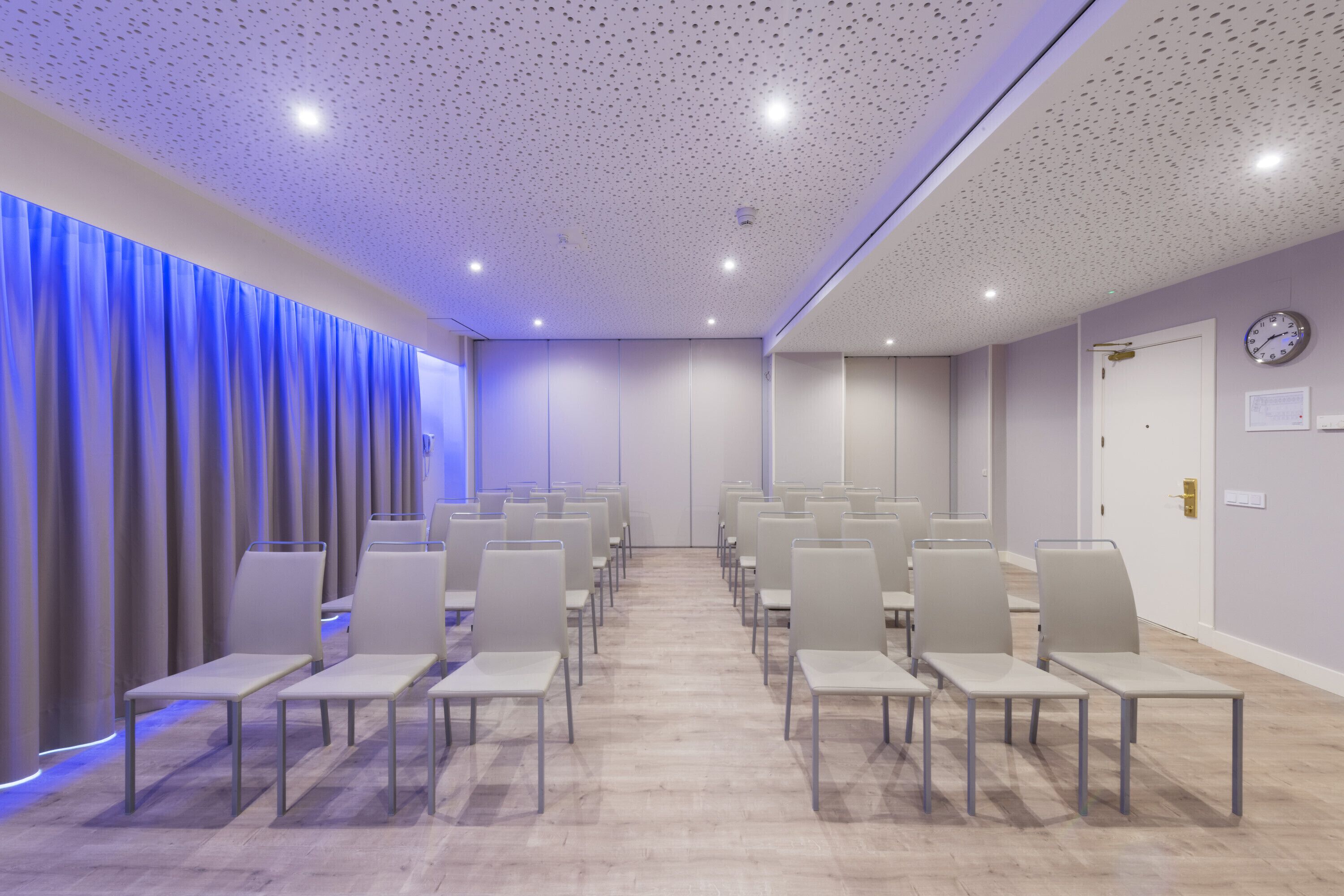 Un salón de conferencias con hileras de sillas, un reloj en la pared y una cortina con luz azul.