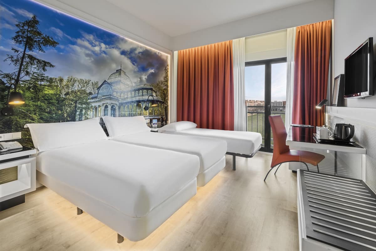 A hotel room with two beds, a desk, a chair, a TV, and a large window with curtains.