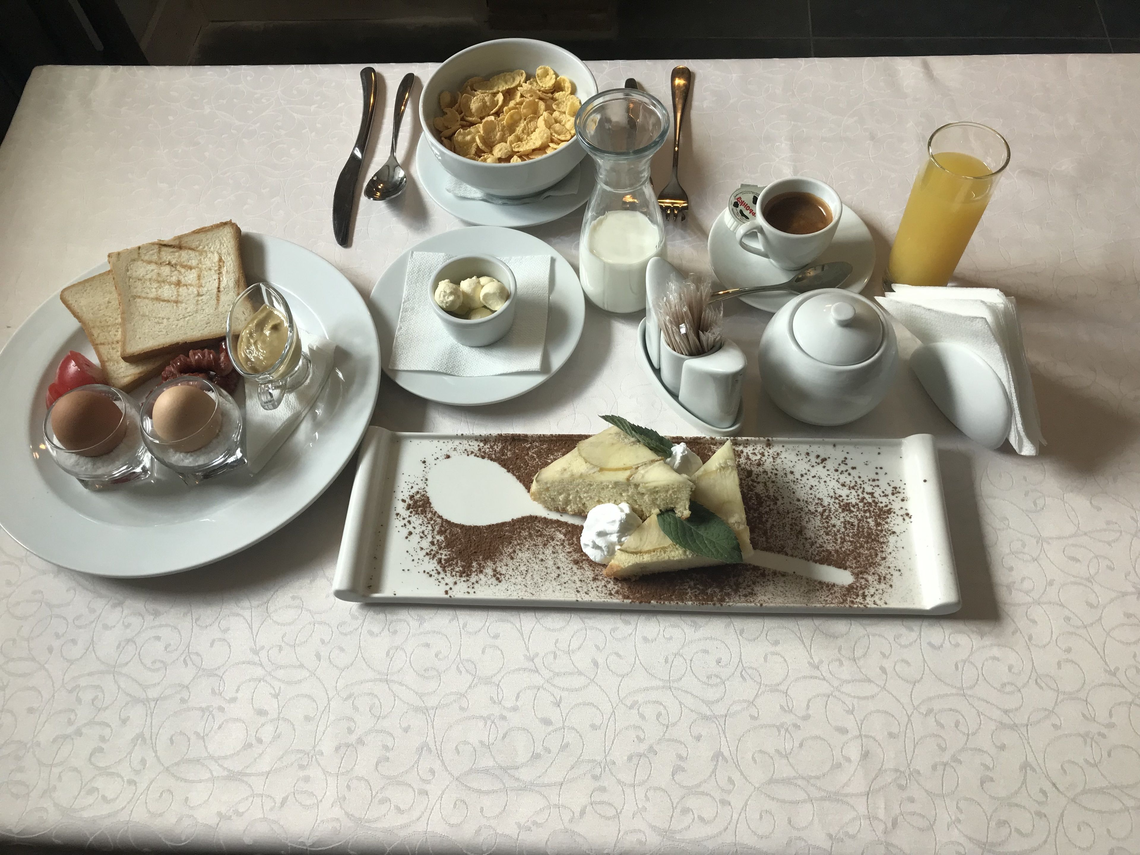 Desayuno con tostadas, huevos, una rebanada de pastel, un vaso de jugo de naranja, un tazón de cereal y una tetera.