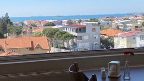 A room with a view of a coastal town, a table with chairs, a vase with flowers, and a window with a view of the sea. - Şakran Otel (Aliaga)