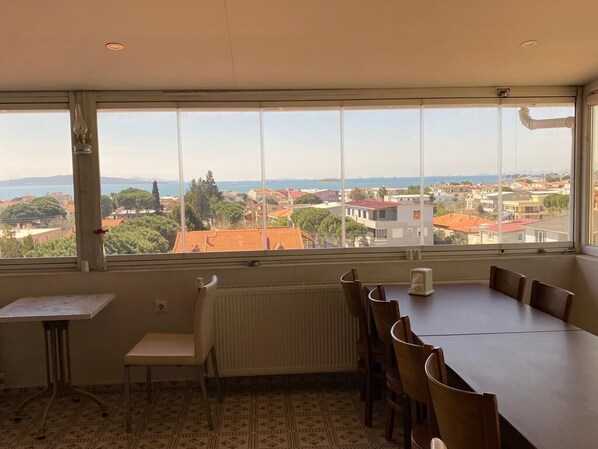 Een kamer met een groot raam dat uitzicht geeft op een kustplaatsje, een eettafel met stoelen, een radiator en een klein tafeltje met een stoel.