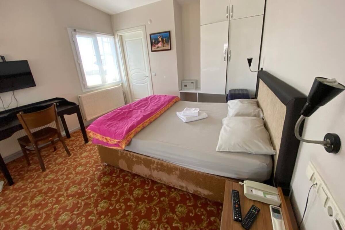 A hotel room with a bed, a desk, a chair, a TV, a wardrobe, and a bedside table with a phone and remote controls.