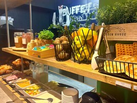 Um buffet de café da manhã com diversas opções de comida, uma máquina de waffles e uma placa com a inscrição 'Little Green House'.