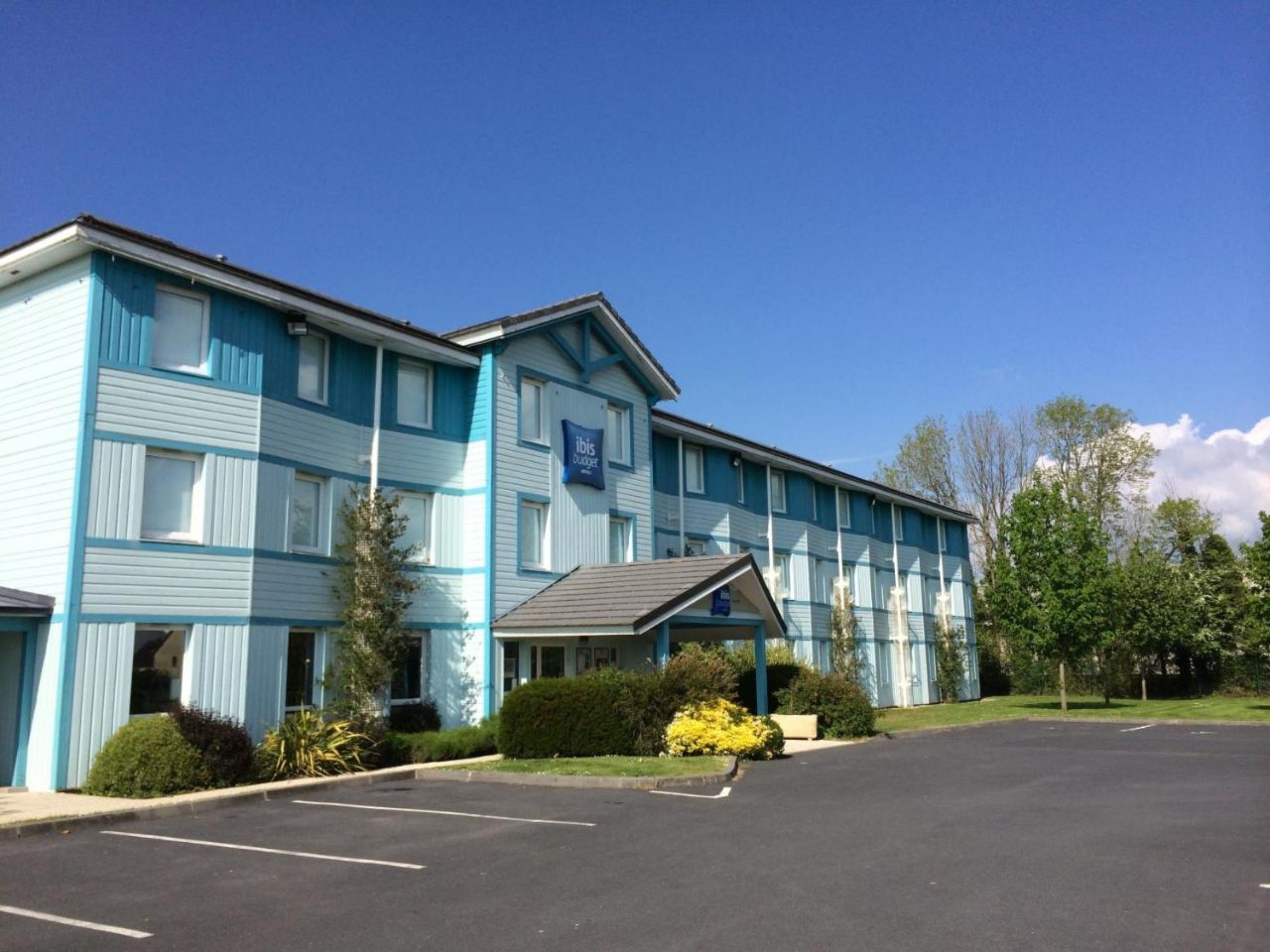 A multi-story hotel with a blue exterior, a parking lot, and a small garden area with yellow flowers.