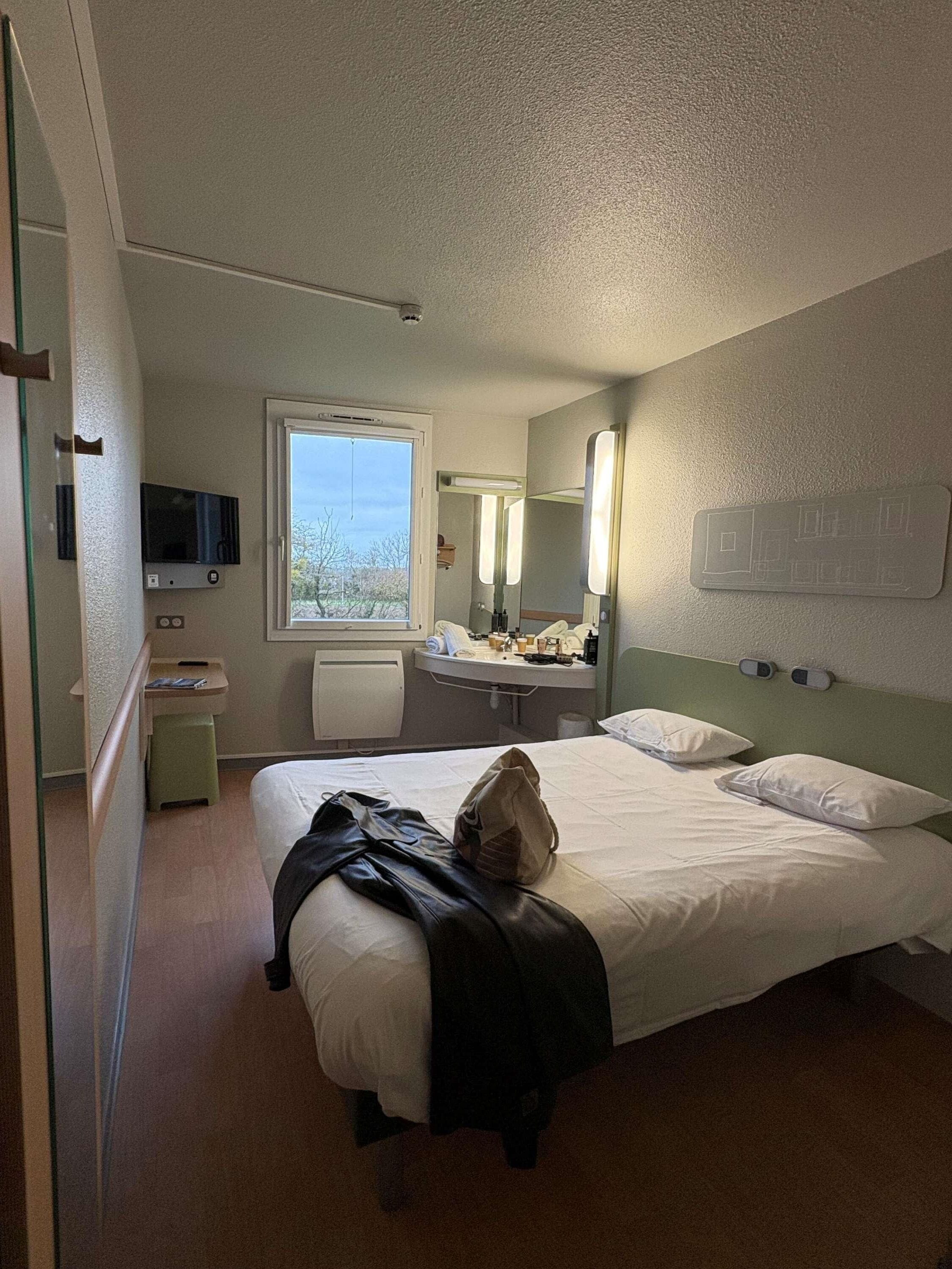 Una habitación de hotel con una cama, un televisor, una ventana, un espejo y una mesita de noche.