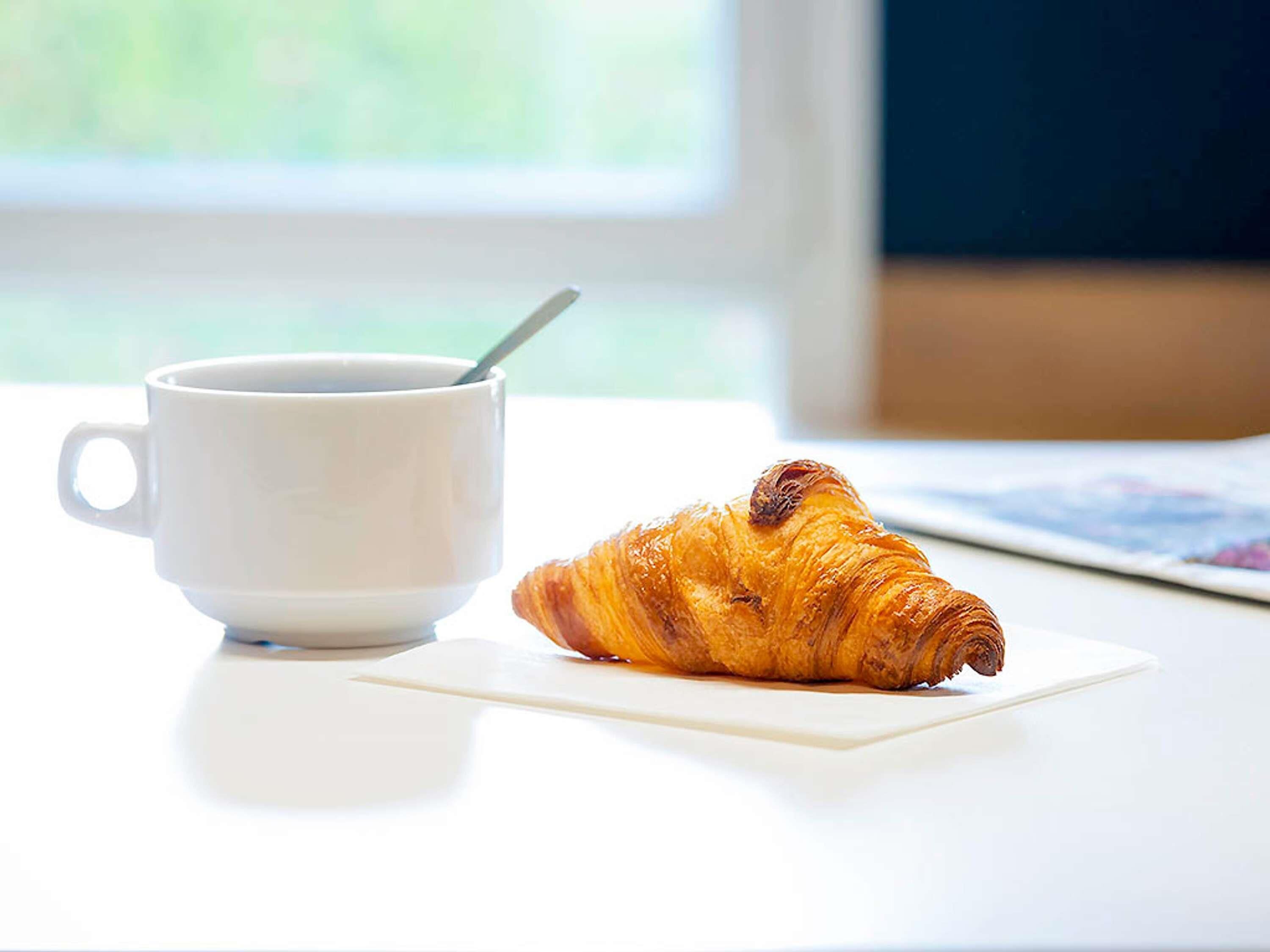A white cup with a spoon on a white table next to a croissant.