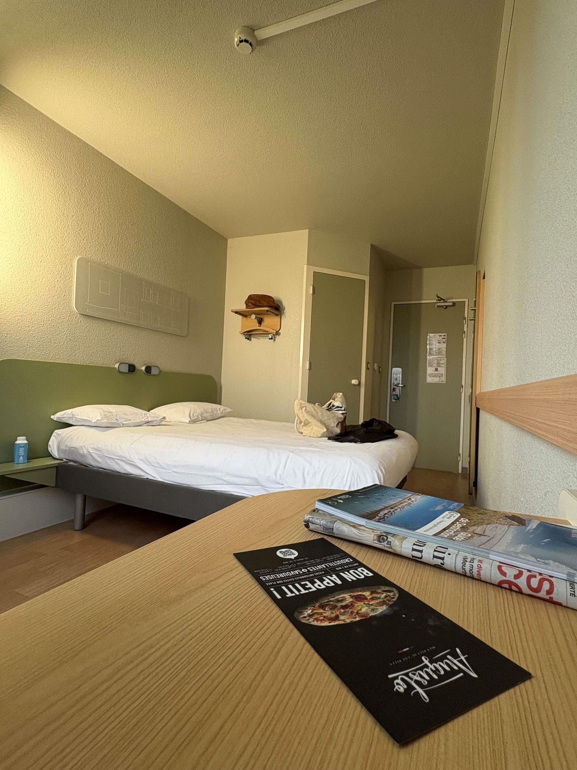 Una habitación de hotel con una cama, una mesa de madera con revistas, una lámpara de pared y una puerta que da a otra habitación.