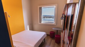 Una cama individual con colchón blanco, una mesita redonda y una ventana que da a una masa de agua y paisaje distante.