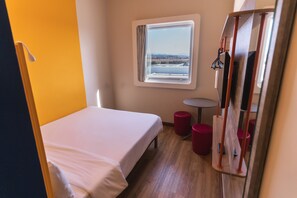Un lit simple avec un matelas blanc, une petite table ronde et une fenêtre donnant sur un plan d’eau et un paysage lointain.