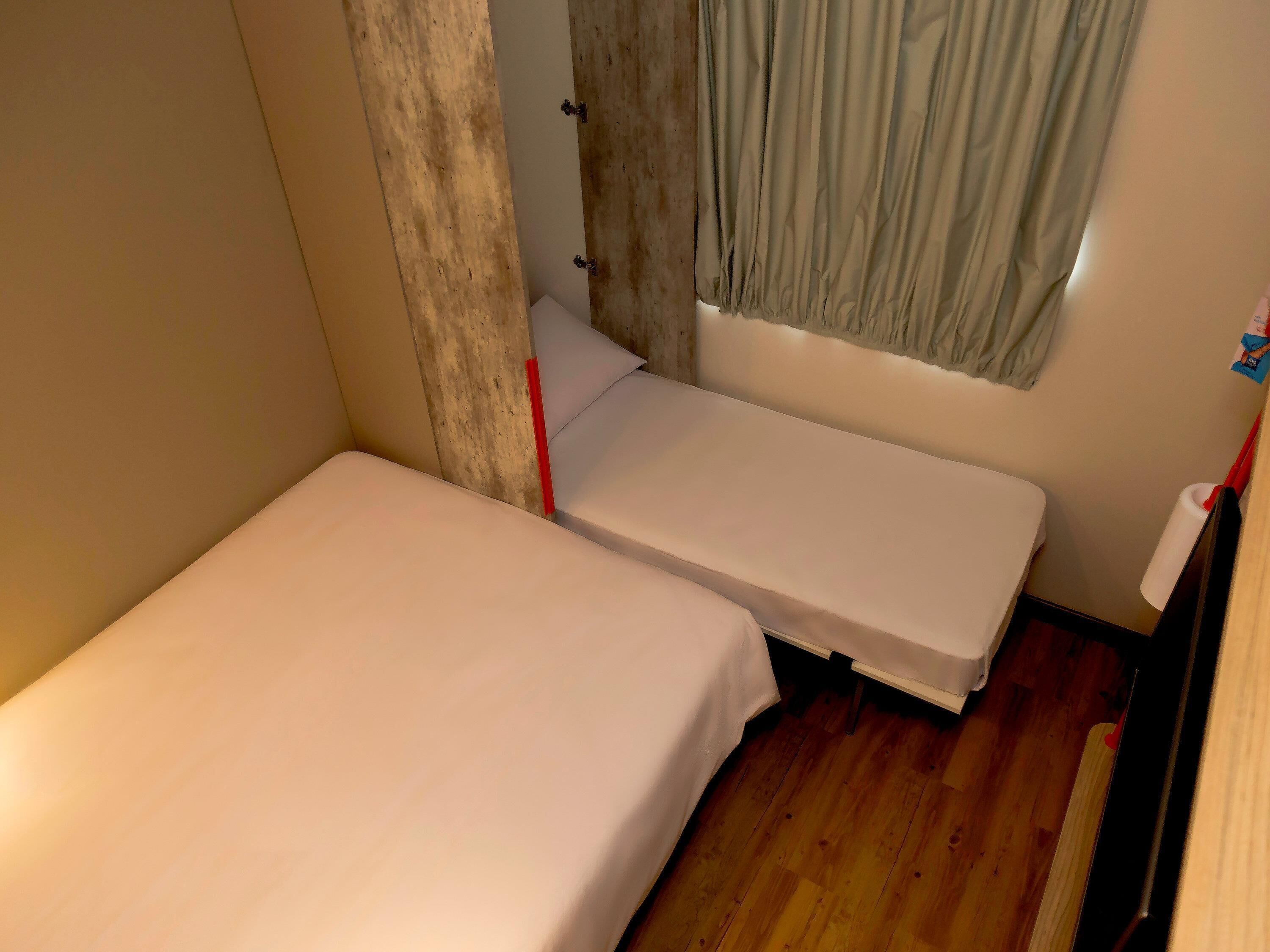 A small, single-bed room with a wooden floor, a window with curtains, and a white wall.