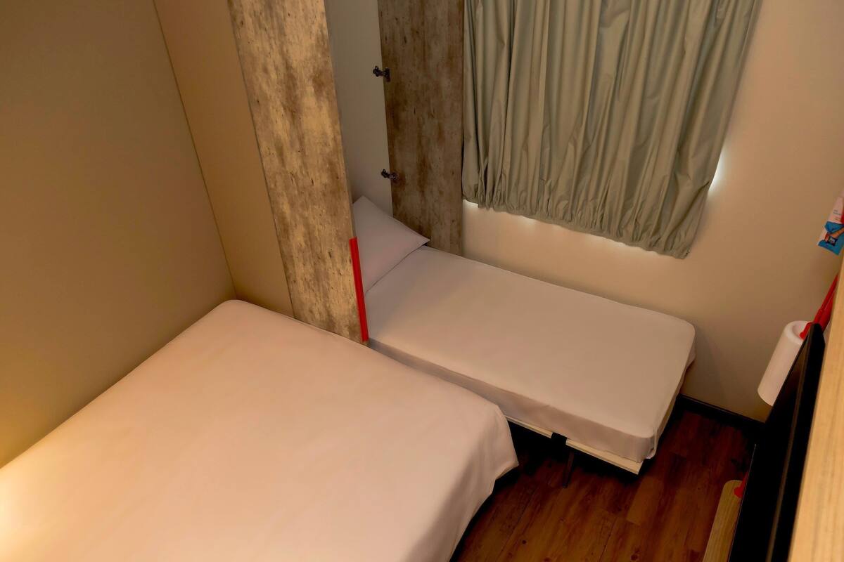 A small, single-bed room with a wooden floor, a window with curtains, and a white wall.