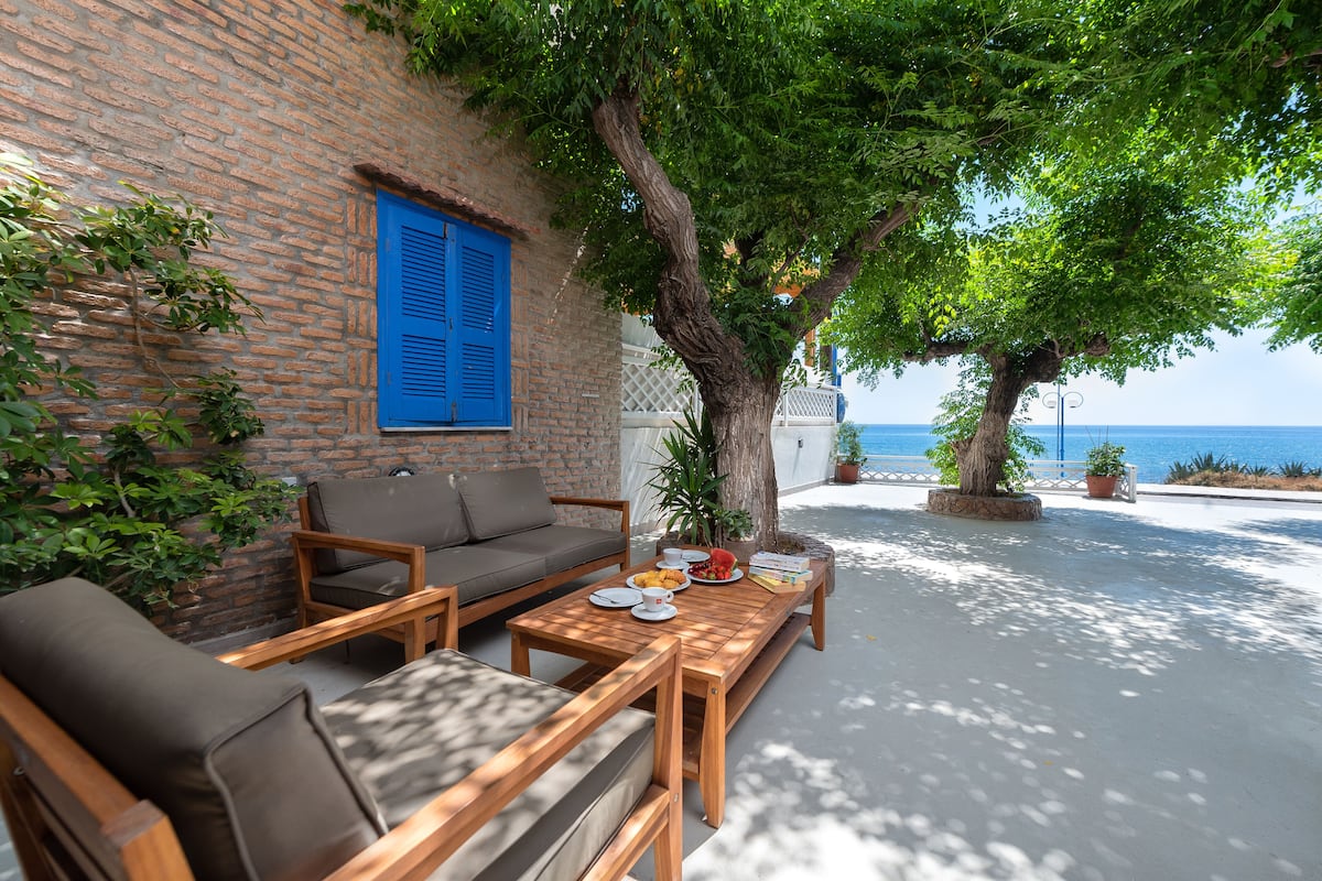 A patio with a wooden table, chairs, and a sofa. There is a brick wall with a blue shutter and a tree providing shade.