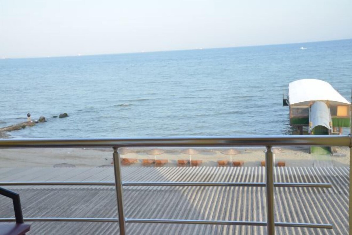 Una terraza con barandilla que da a un mar tranquilo, un pequeño bote fondeado cerca y una playa de arena al fondo.