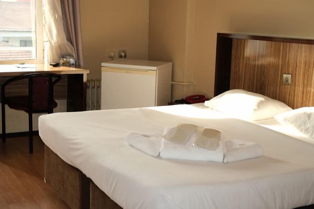 Habitación de hotel con una cama bien hecha, un escritorio con silla, un refrigerador pequeño y un ventanal con cortinas.