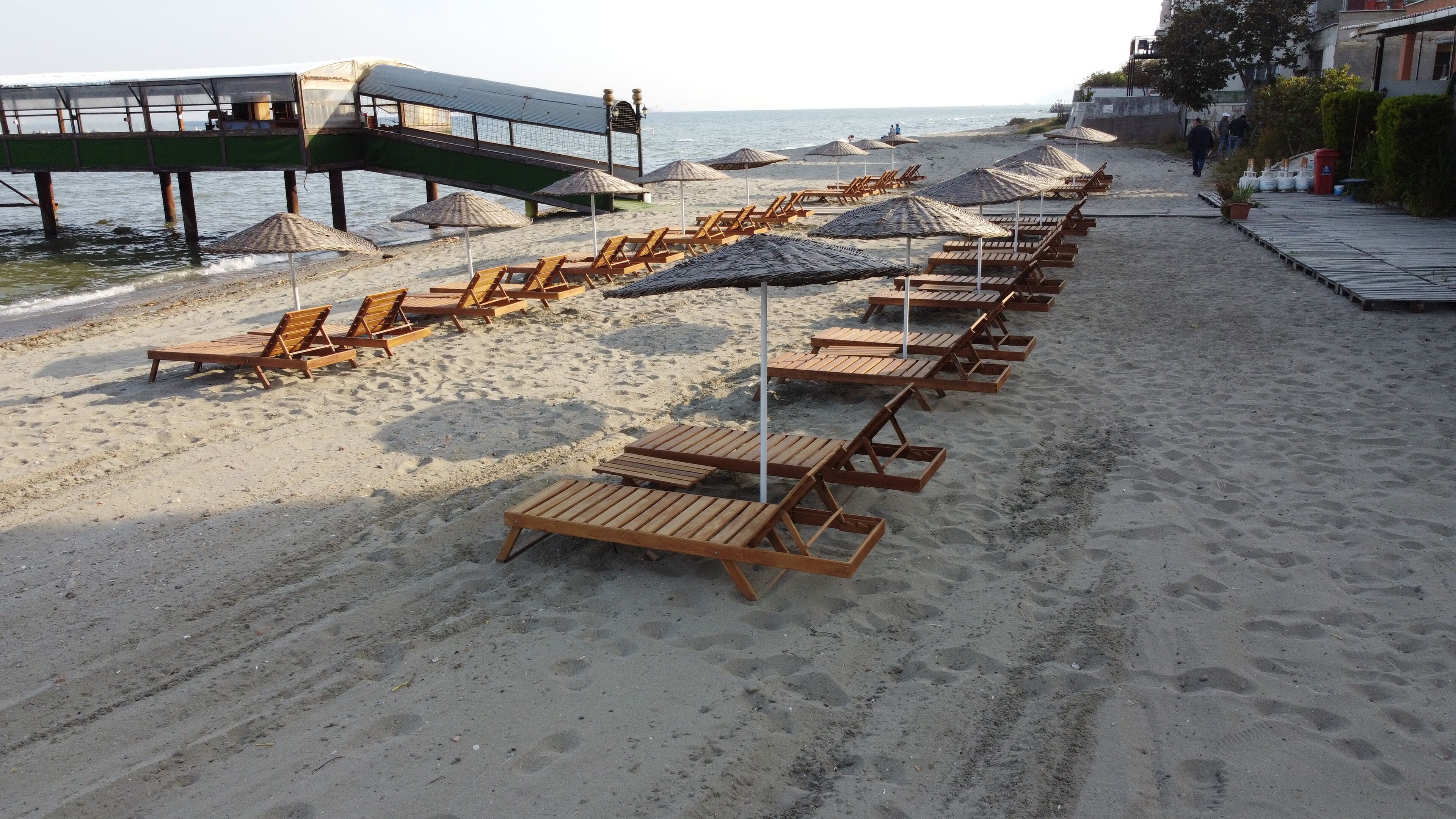 A beach with rows of wooden sun loungers and thatched umbrellas, a walkway leading to a pier, and the sea in the background.