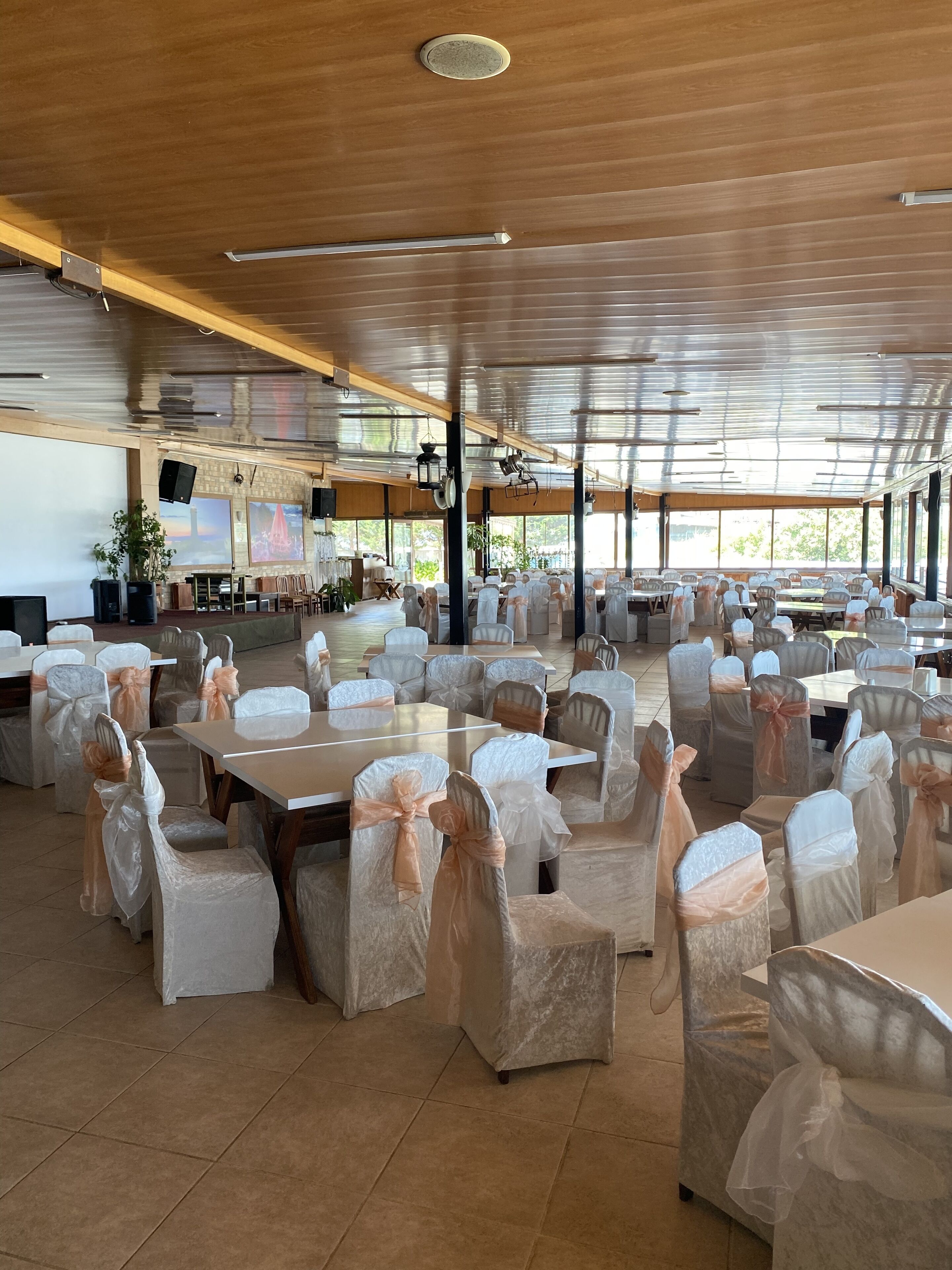 A spacious indoor venue with multiple tables and chairs, some covered with white and peach-colored chair covers, and a high ceiling with wooden beams and a skylight.