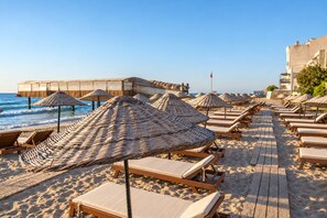 Una playa con sillones de madera y sombrillas tejidas, un paseo marítimo de madera y un edificio con una bandera.