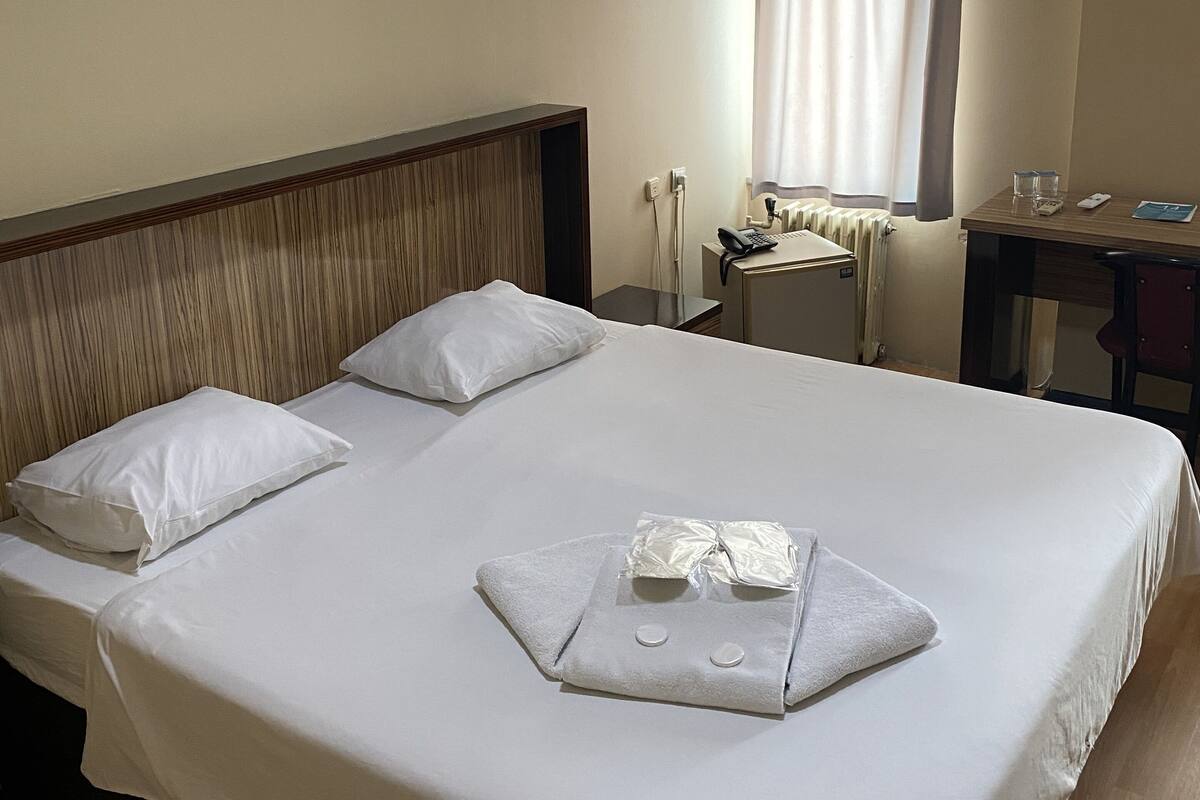 A hotel room with a neatly made bed, a desk with a chair, a window with curtains, and an air conditioning unit on the wall.