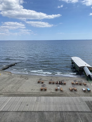 Una playa con sillones de madera y sombrillas, una terraza de madera, y un muelle que se adentra en el agua.