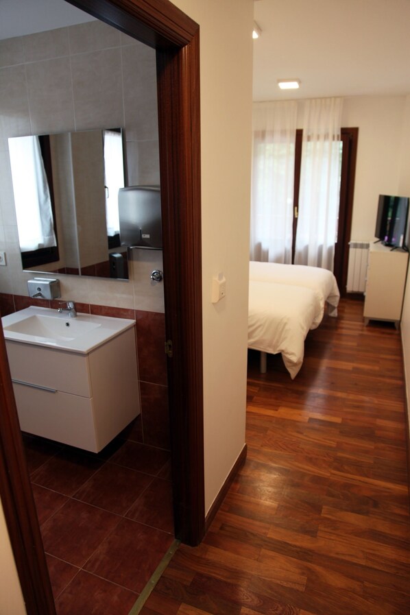 A hotel room with a wooden floor, a bed, a small desk, a television, and a bathroom with a sink and mirror.
