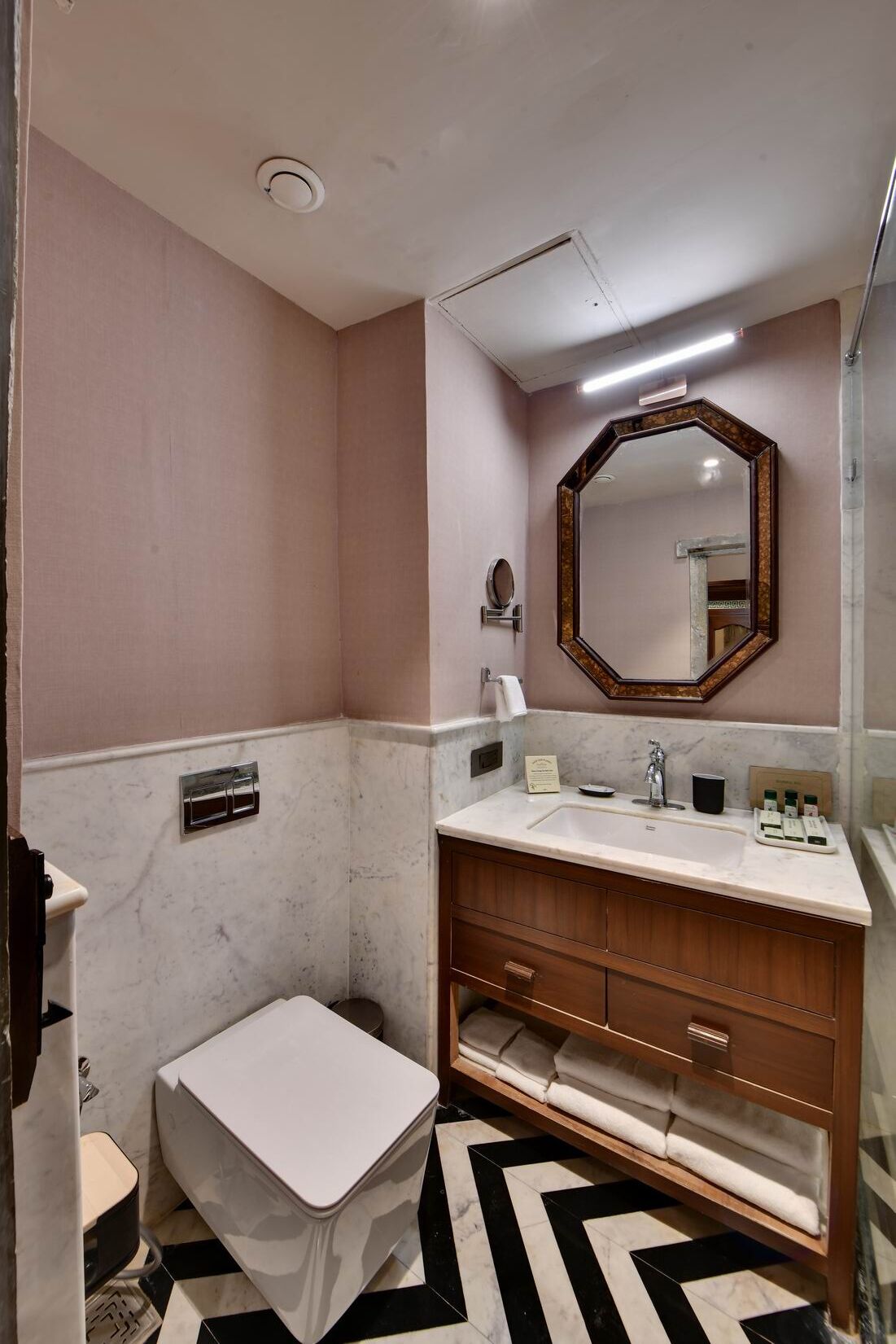 Une salle de bain avec des toilettes blanches, un meuble-lavabo en bois doté d’un miroir et un sol à motifs noir et blanc.