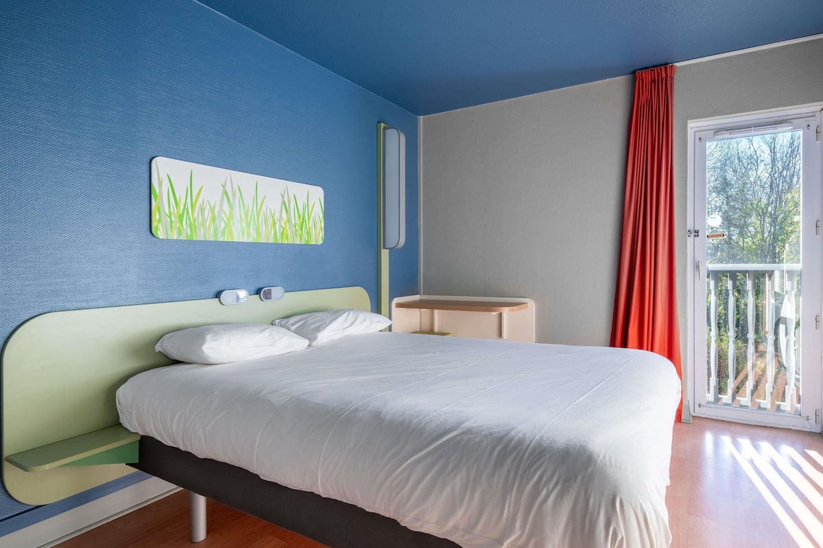 Une chambre avec un lit, une table de chevet, une lampe fixée au mur, une fenêtre avec un rideau rouge et une vue sur la verdure à l’extérieur.