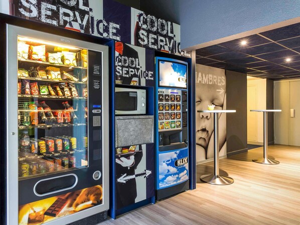 Máquina de vendas com variedade de lanches e bebidas, micro-ondas e área de serviço com placa indicando 'COOL SERVICE'.