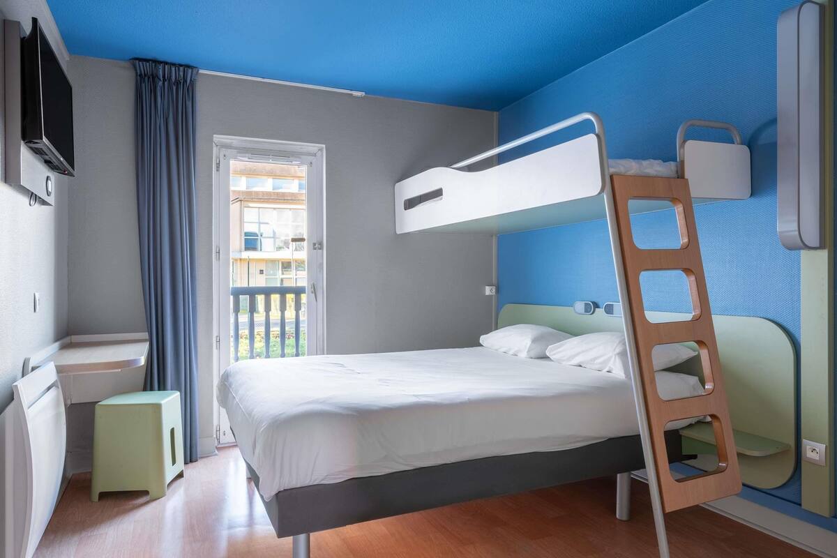 A bunk bed with a ladder, a small stool, a television, and a window with curtains.