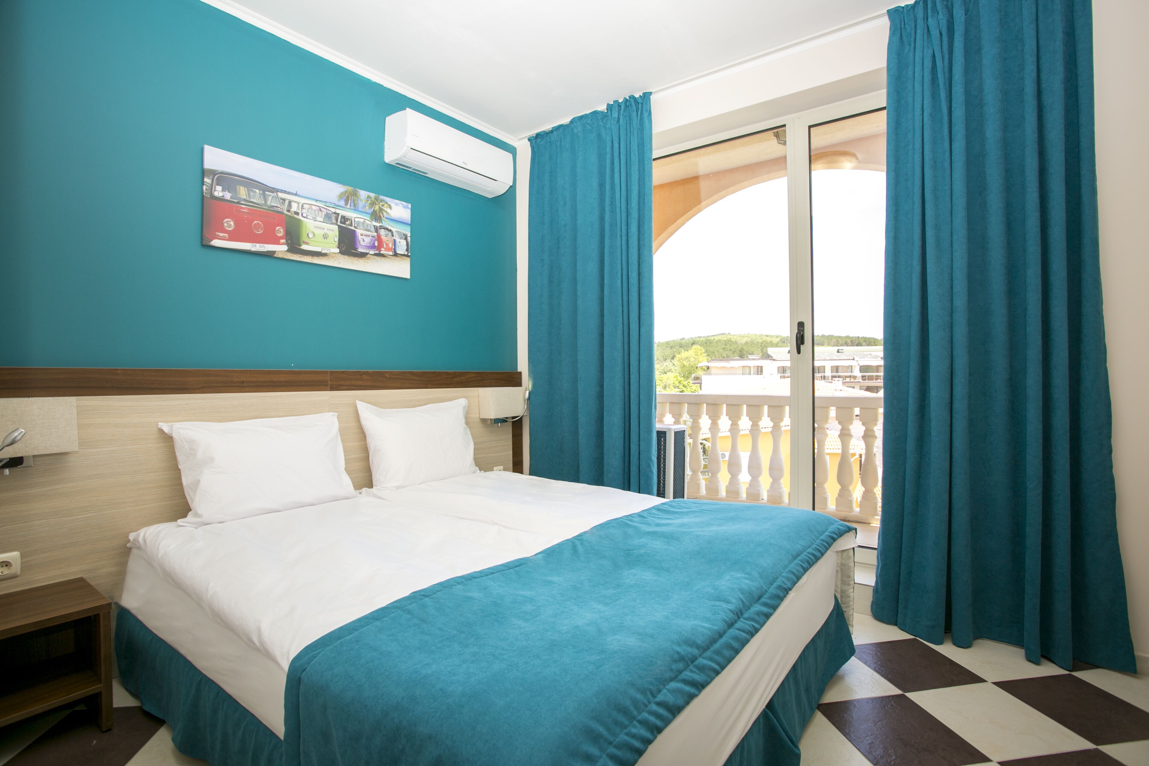 A hotel room with a bed, a window with curtains, a wall-mounted air conditioner, and a painting of buses.