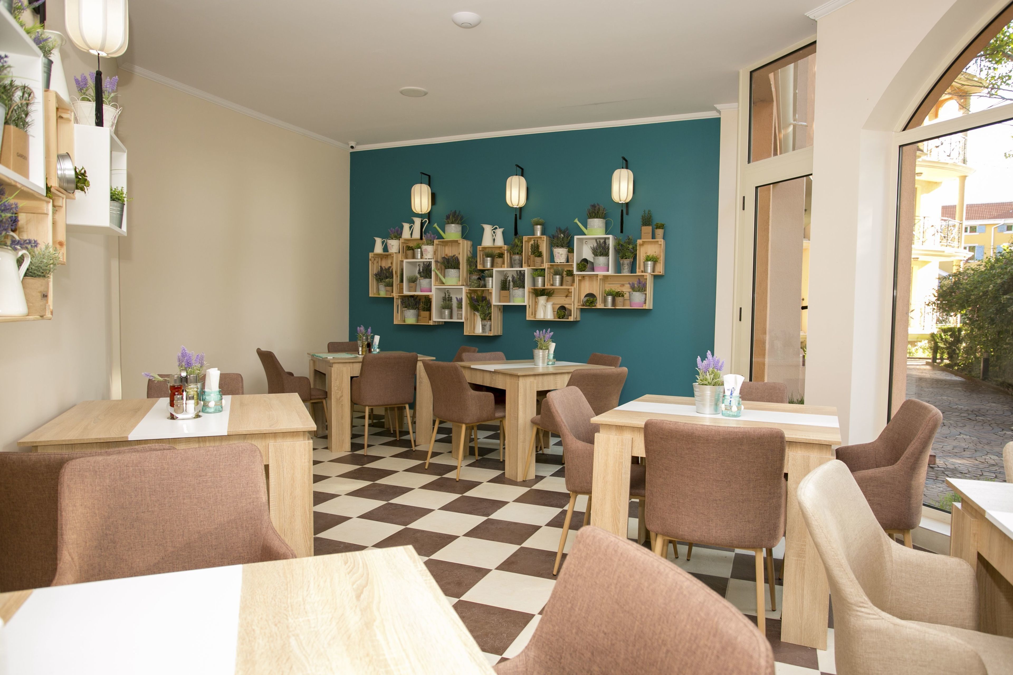Un café con mesas y sillas de madera, suelo a cuadros y una pared decorada con estanterías de madera que sostienen plantas e iluminación.