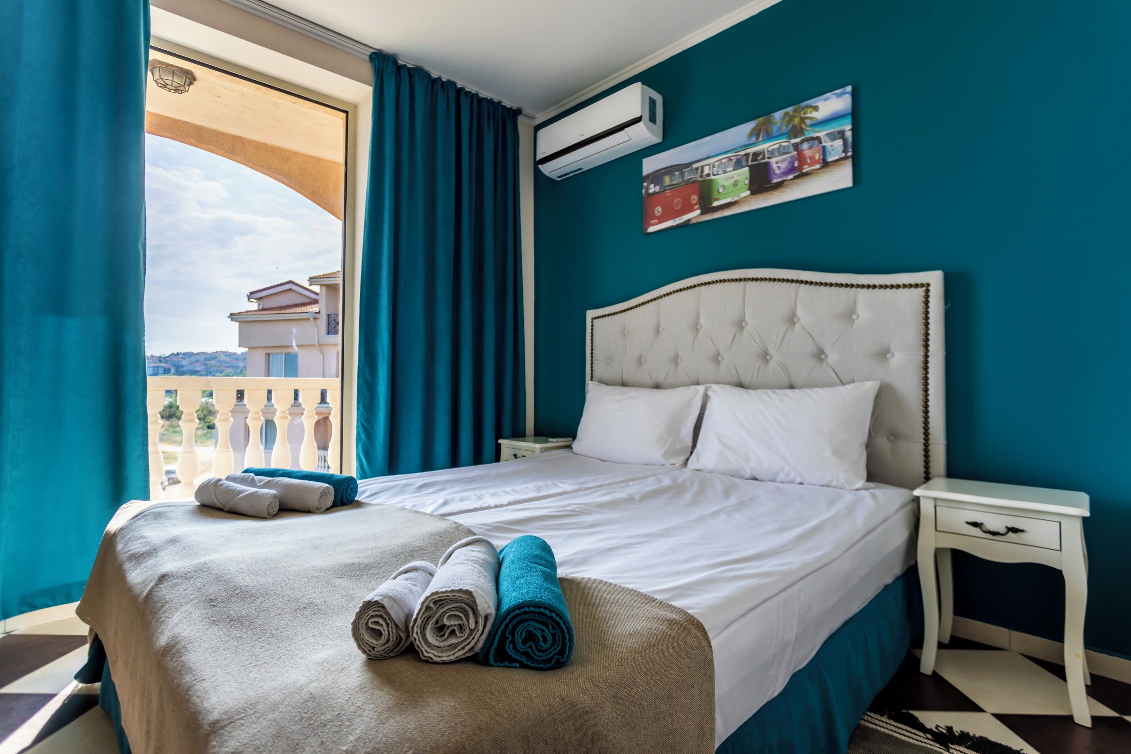 A hotel room with a bed, a nightstand, a window with curtains, an air conditioning unit, and a wall-mounted picture.