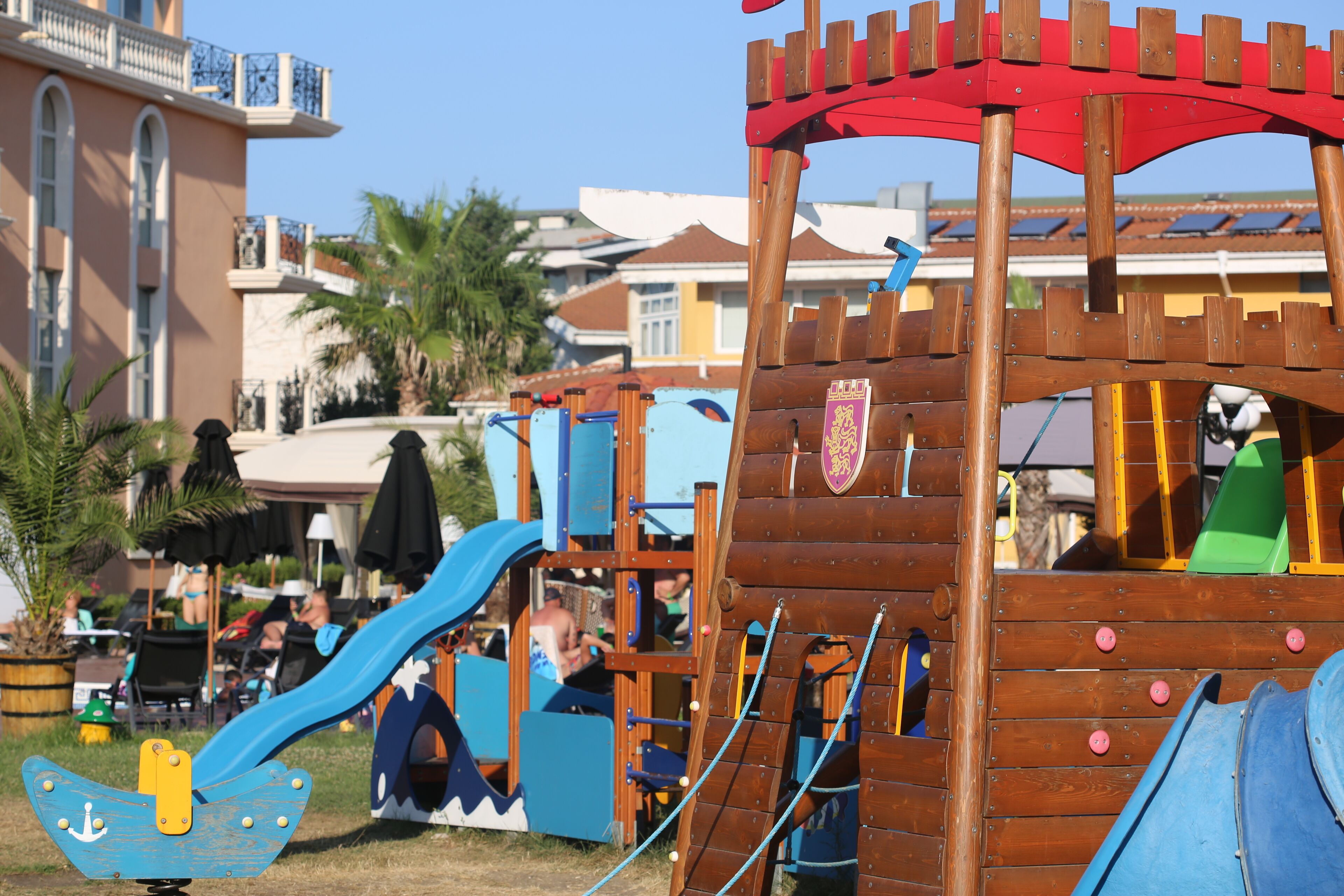 A wooden play structure with slides and climbing elements, surrounded by palm trees and a building with balconies.