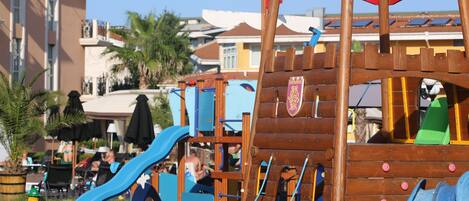A wooden play structure with slides and climbing elements, surrounded by palm trees and a building with balconies.