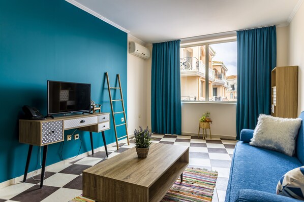 A modern living room with a checkered floor, a blue sofa, a wooden coffee table, a desk with a TV, and a view of a balcony with blue curtains. - Hacienda Beach Resort (Sozopol)