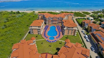 A large hotel with a central swimming pool surrounded by lounge chairs, a beach view, and a parking area.