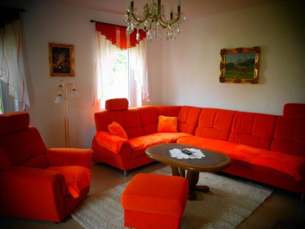 A living room with a large orange sectional sofa, a small orange armchair, and a round coffee table. There is a chandelier, a floor lamp, and two paintings on the walls.