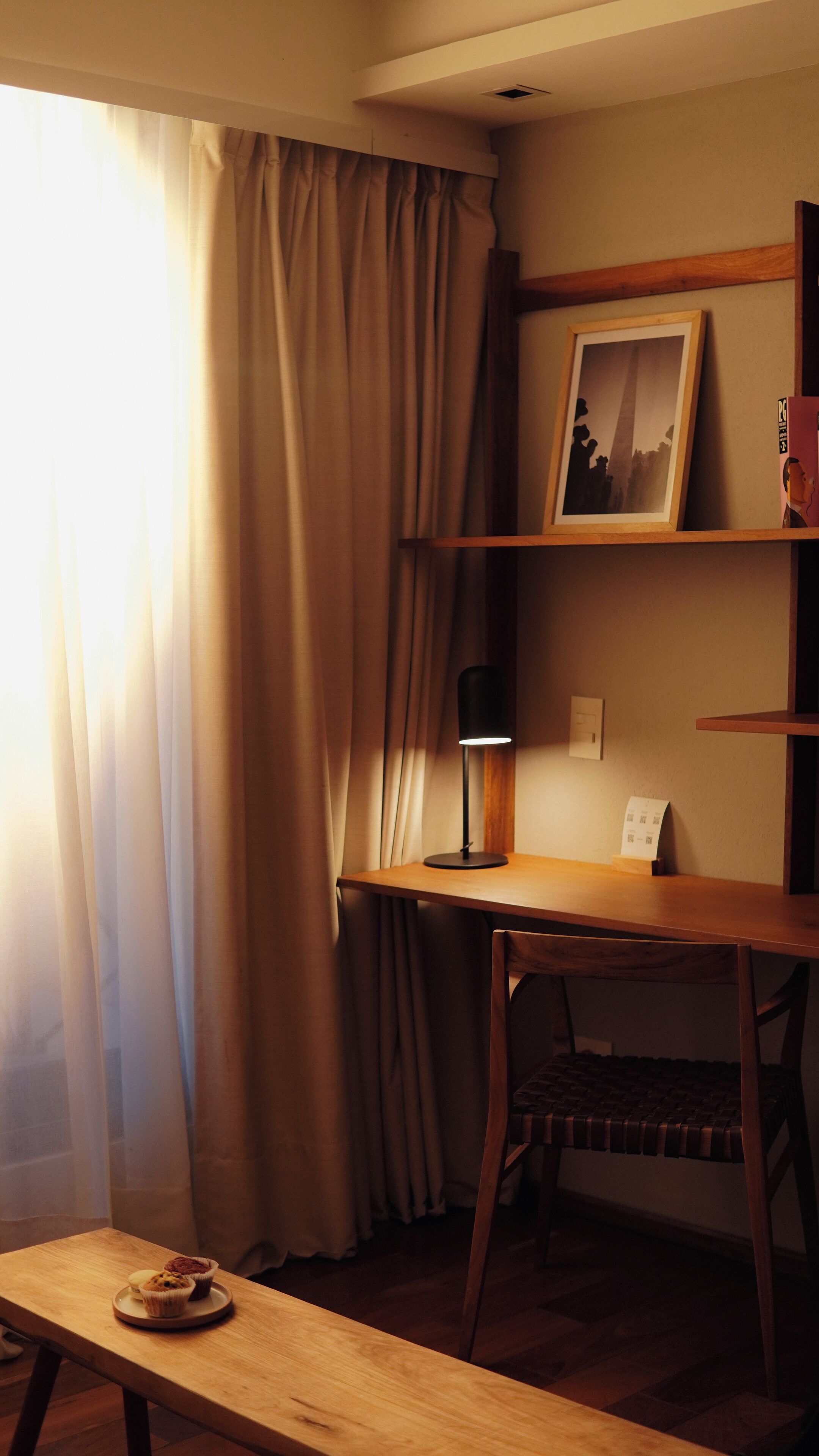 A room with a wooden desk, a chair, a lamp, a framed picture, and a window with curtains.