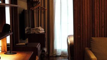 Habitación de hotel pequeña con piso de madera, un escritorio con silla, un sofá, un televisor y una ventana con cortinas.