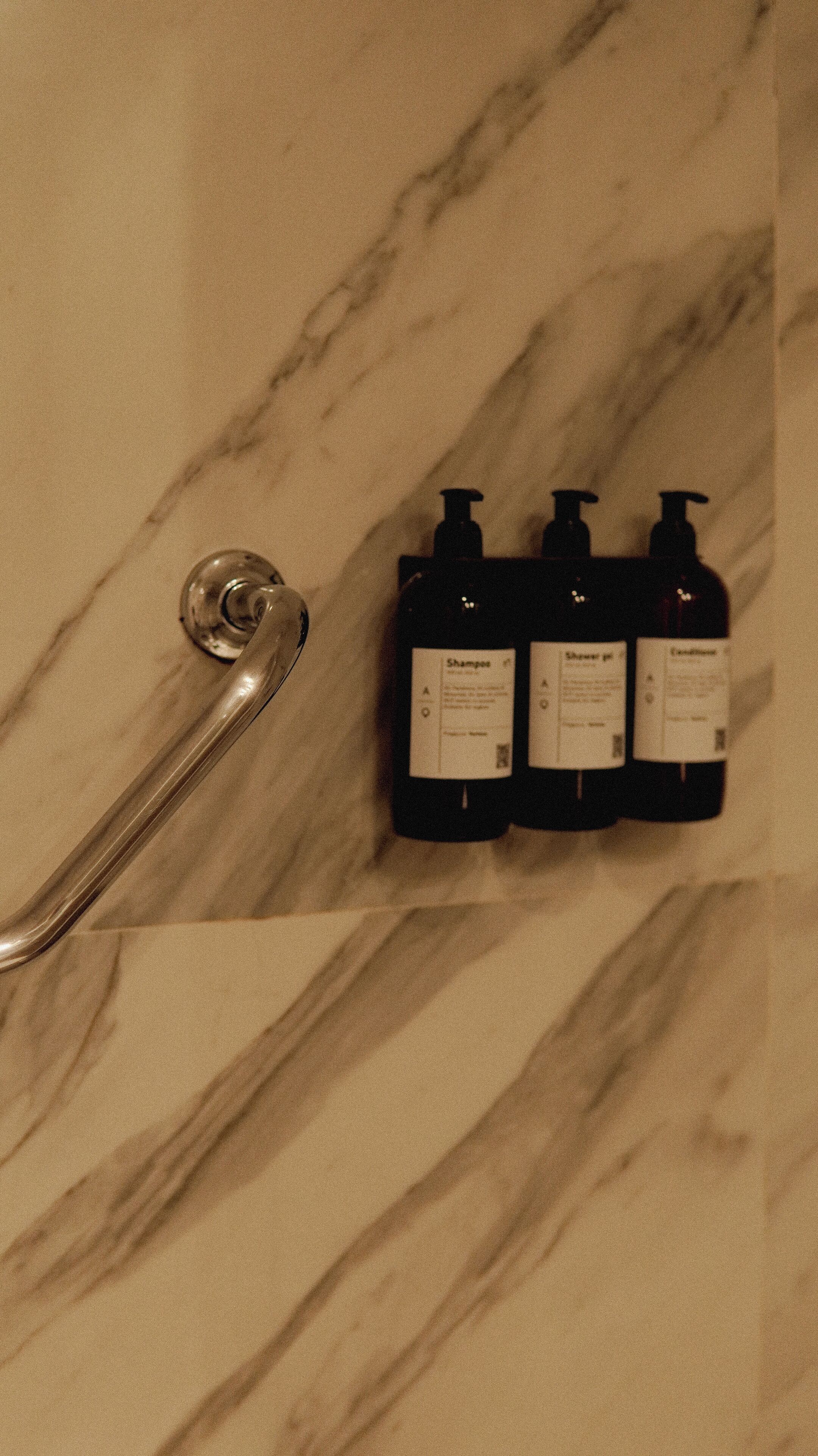 Un baño con piso de mármol, un grifo de plata y tres botellas negras con la etiqueta 'Shampoo' en un estante.