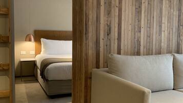 A bedroom with a wooden panel wall, a bed with white bedding, a beige sofa, and a nightstand with a lamp.